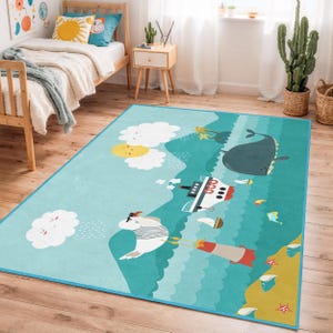 Puede incluir: Una alfombra rectangular azul claro para niños con temática náutica. La alfombra presenta ilustraciones de una ballena, un barco, nubes, sol y una gaviota con gorro de marinero. La alfombra tiene un borde azul y está colocada sobre un suelo de madera.