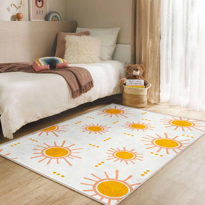 May include: A rectangular white rug with a repeating pattern of orange suns and pink rays. The rug is placed on a wooden floor in a bedroom setting, with a bed and decorative items in the background.