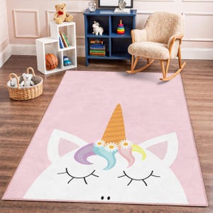 Può includere: Un tappeto rettangolare rosa con un grande volto di unicorno. L'unicorno ha gli occhi chiusi, un corno arancione a strisce e una corona floreale. Il tappeto è progettato per la cameretta dei bambini.