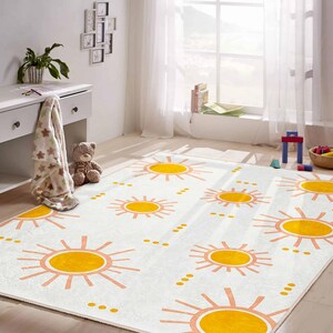 May include: A rectangular white rug with a repeating pattern of yellow suns and peach-colored rays. The rug is in a child's room, with a white desk, a teddy bear, and a window with sheer curtains in the background.