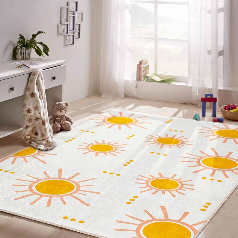 May include: A rectangular white rug with a repeating pattern of yellow suns and peach-colored rays. The rug is in a child's room, with a white desk, a teddy bear, and a window with sheer curtains in the background.