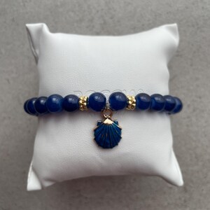 Könnte beinhalten: Ein elastisches Armband mit glatten, runden, dunkelblauen Perlen, die mit kleinen, texturierten goldfarbenen Metallzwischenstücken abwechseln. Ein goldfarbener Metallanhänger in Form einer Muschel mit blauen Emaille-Details hängt in der Mitte.