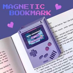 May include: A purple magnetic bookmark in the shape of a retro handheld game console. The screen shows "SAVE PROGRESS?" with "yes" and "One more chapter" options. The text "MAGNETIC BOOKMARK" is at the top. The bookmark is on a book page.