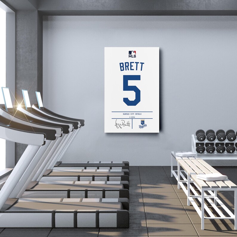 May include: A framed white canvas featuring the name "BRETT" and the number "5" in blue, with the MLB logo. Below, it reads "Kansas City Royals" and includes a signature. The artwork is displayed in a gym setting.