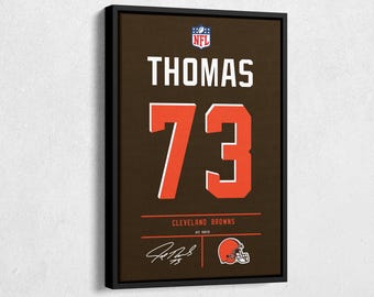 May include: Framed artwork featuring a brown background with the name "THOMAS" and the number "73" in orange and white. The NFL logo is at the top, with "CLEVELAND BROWNS" and a signature at the bottom. A football helmet graphic is also included.