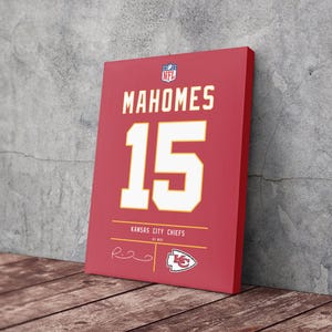 May include: A red canvas print featuring the name "MAHOMES" and the number "15" in white. The print also includes "KANSAS CITY CHIEFS" and a team logo. The NFL logo is visible in the top corner. The canvas is leaning against a gray wall on a wooden surface.