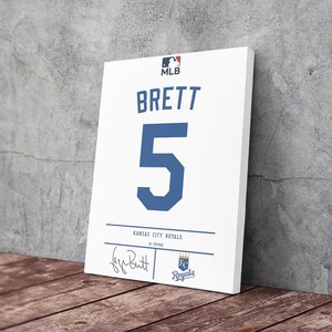 May include: White canvas art print of a baseball jersey. The jersey features the name "BRETT" and the number "5" in blue. The MLB logo is at the top, with "Kansas City Royals" and a signature at the bottom.