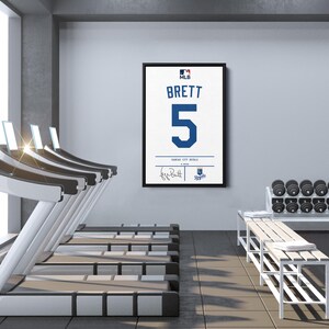 May include: Framed white baseball jersey with the name "BRETT" and the number "5" in blue. The jersey is from the Kansas City Royals, with a signature and team logo. The framed jersey hangs on a gray wall in a gym setting.