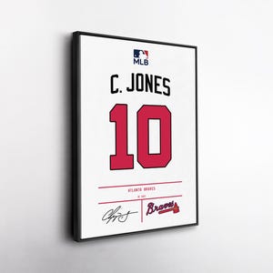 May include: Framed white jersey with the name "C. JONES" and the number "10" in red. The MLB logo is at the top, with "Atlanta Braves" and a signature at the bottom. The jersey is in a black frame.
