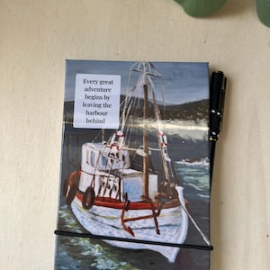 Può includere: Un piccolo taccuino con un dipinto di una barca da pesca bianca e rossa sull'acqua. Il testo "Every great adventure begins by leaving the harbour behind" è sul davanti. Una penna nera è accanto al taccuino.