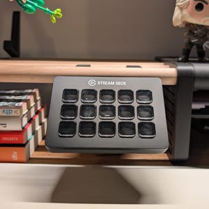 Könnte beinhalten: Ein schwarzes Stream Deck mit 15 programmierbaren Tasten liegt auf einem Holzregal. Das Gerät hat das Stream Deck-Logo und ist von anderen Gegenständen umgeben, darunter Bücher und eine Figur.