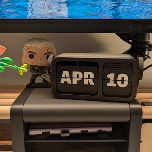 Könnte beinhalten: Ein schwarzer Klappkalender mit der Anzeige "APR 10" in Weiß. Der Kalender steht auf einem schwarzen Monitorständer mit einem Regal, das ein lila Gerät enthält. Eine Figur eines Charakters mit weißem Haar und Bart ist sichtbar.