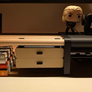Könnte beinhalten: Ein hellfarbener Schreibtisch-Organizer mit zwei Schubladen und schwarzen Griffen. Eine kleine Figur eines Charakters mit einem Schwert steht auf dem Schreibtisch. Links ist ein Stapel Bücher zu sehen.