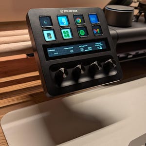 Könnte beinhalten: Ein schwarzes Stream Deck Bedienfeld mit mehreren Tasten und einem kleinen Display. Die Tasten zeigen App-Symbole, und es gibt vier Drehregler unter dem Display. Das Gerät ist für die Erstellung von Inhalten und Streaming konzipiert.