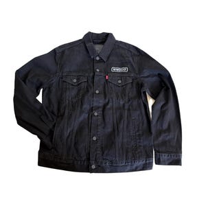 Puede incluir: Chaqueta vaquera negra con cierre de botones, dos bolsillos en el pecho y bolsillos laterales. La chaqueta presenta un pequeño parche blanco con "WWDC17" en el pecho izquierdo y una etiqueta roja en el bolsillo izquierdo del pecho.