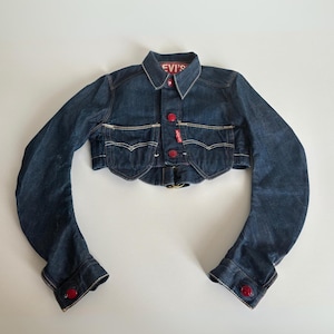 Op de afbeelding: Een donkerblauwe, korte spijkerjas met lange mouwen. Het heeft rode knopen en witte stiksels. Het Levi's-logo staat op de kraag en een klein label. De jas heeft twee voorzakken. De stijl van de jas is cropped.