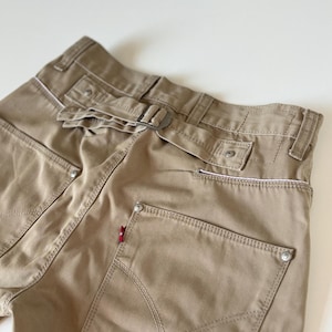 Könnte beinhalten: Khakifarbene Hose mit Gürtel und Metallschnalle. Die Gesäßtaschen haben eine einzigartige Form und Nahtdetails. Ein kleines rotes Etikett ist auf einer der Taschen sichtbar. Die Hose ist aus robustem Stoff gefertigt.