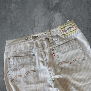 Könnte beinhalten: Nahaufnahme einer beige-weiß gestreiften Levi Strauss & Co. Jeans. Die Jeans hat ein rotes Levi's-Etikett, braune Nähte und ein Two Horse Brand-Etikett. Die Gesäßtaschen haben ein markantes geschwungenes Design.