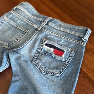 Peut inclure: Jean bleu clair avec une poche arrière effilochée ornée d'un écusson rouge, blanc et bleu marine. Le jean présente une coupe classique à cinq poches et un délavage.