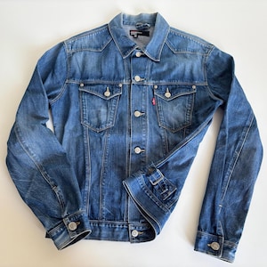 Puede incluir: Chaqueta vaquera azul con un diseño clásico. Cuenta con cierre de botones, dos bolsillos en el pecho y dos bolsillos laterales. La chaqueta tiene un lavado descolorido y una pequeña etiqueta roja en el bolsillo del pecho.