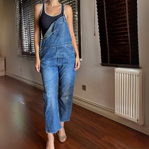 Può includere: Salopette in denim blu con pettorina e spalline regolabili. La salopette ha una vestibilità rilassata, dettagli strappati e un orlo grezzo. Sotto è indossata una canotta nera con rifiniture bianche. Stile vintage.