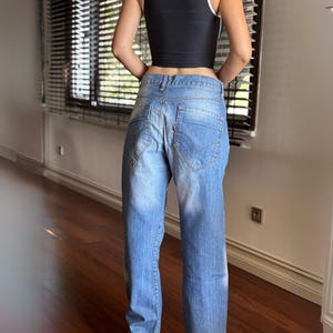 May include: Light blue denim jeans with a faded wash and a red logo patch on the back pocket. The jeans have a classic five-pocket design and a relaxed fit. The model is wearing a black vest top.