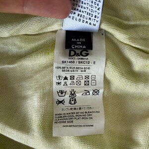 May include: A close-up of a clothing tag with the text "MADE IN CHINA" and the D&G logo. The tag also includes washing instructions and the text "100% SETA-SILK-SEDA-SOIE". The background is a light green fabric.