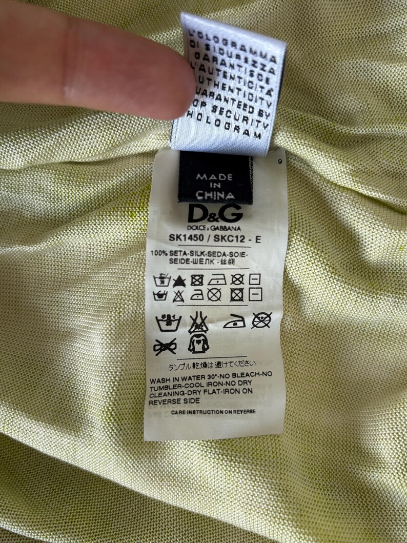 May include: A close-up of a clothing tag with the text "MADE IN CHINA" and the D&G logo. The tag also includes washing instructions and the text "100% SETA-SILK-SEDA-SOIE". The background is a light green fabric.