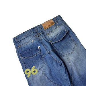 May include: Faded blue denim jeans with multiple pockets and decorative stitching. The jeans feature a light brown leather patch and a yellow embroidered "96" on the back pocket. The jeans have a relaxed fit.