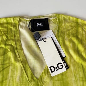 May include: A lime green V-neck sweater with a tie-dye effect. The sweater has a tag with the D&G logo attached. The tag is white with black text. The sweater is made of a lightweight material.
