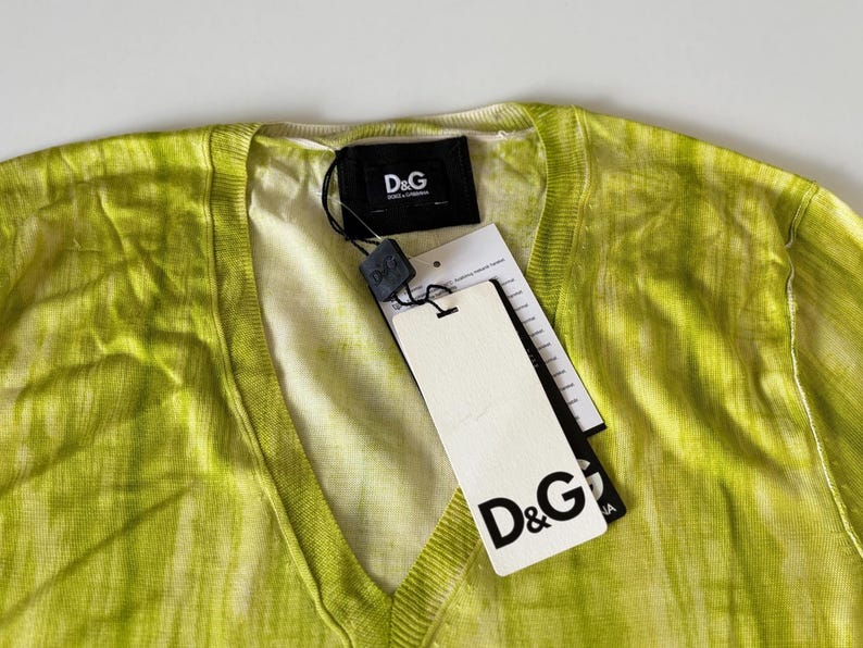 May include: A lime green V-neck sweater with a tie-dye effect. The sweater has a tag with the D&G logo attached. The tag is white with black text. The sweater is made of a lightweight material.