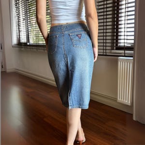 May include: A light blue denim skirt with a back slit. The skirt has two back pockets and a small logo on the right pocket. The skirt is knee-length and has a classic, casual style.