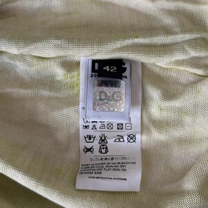May include: Close-up of a garment tag with the brand logo "D&G" and size 42. The tag includes care instructions with symbols and text, such as "Wash in water 30&deg; - No bleach - No tumbler". The background is a light green fabric.