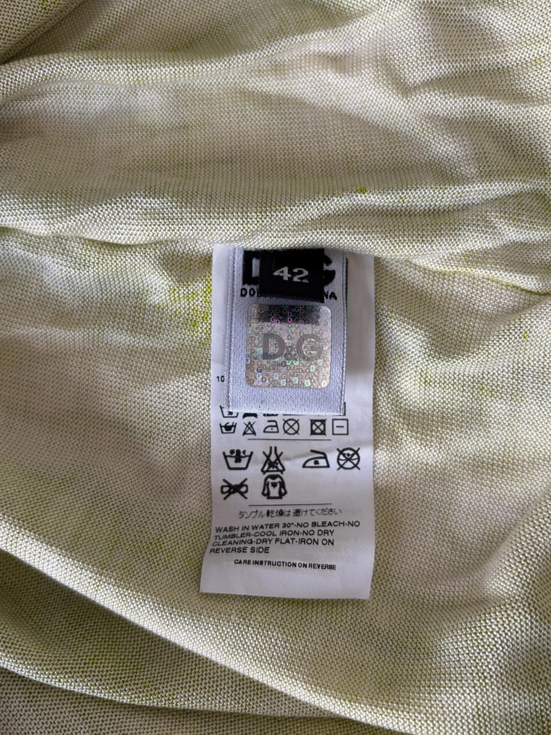 May include: Close-up of a garment tag with the brand logo "D&G" and size 42. The tag includes care instructions with symbols and text, such as "Wash in water 30&deg; - No bleach - No tumbler". The background is a light green fabric.