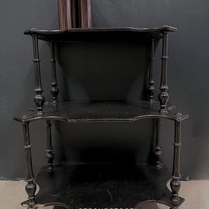 Peut inclure: Étagère antique noire à trois niveaux avec pieds décoratifs et bords festonnés. Deux bouteilles marron avec une plante sont sur l'étagère supérieure. L'étagère est contre un mur gris foncé. Le texte "@SECONDSTOORI" est en bas.