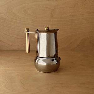 May include: A polished stainless steel moka pot featuring a rounded base and a cylindrical upper chamber. The coffee maker includes a vertical handle and a lid knob, both finished in a bright, metallic gold tone, providing a distinct contrast.