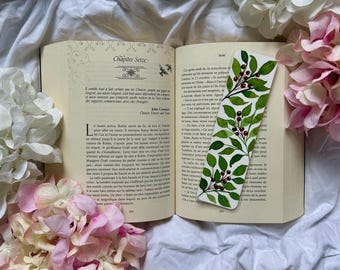 May include: An open book with a decorative bookmark featuring green leaves and red berries. The bookmark is placed between the pages of a book with French text. Pink and white flowers are arranged around the book on a white fabric surface.