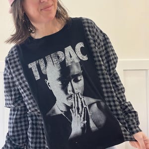 May include: Black graphic t-shirt featuring a black and white image of Tupac Shakur with the word "TUPAC" in white block letters. The shirt has long sleeves with a black and gray plaid pattern.