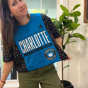 Puede incluir: Una camiseta azul con el texto "CHARLOTTE FOOTBALL CLUB" en blanco. La camiseta tiene el logo de Adidas y un emblema circular. La persona de la imagen lleva una camiseta negra con estampado floral y pantalones cargo verde oliva.