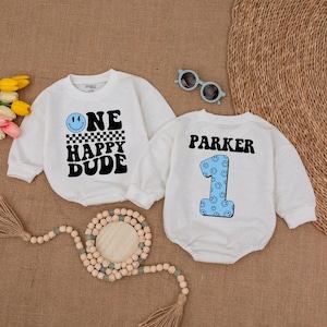 May include: Two white baby rompers with long sleeves. One has the text "ONE HAPPY DUDE" with a smiley face and checkered pattern. The other has the name "PARKER" and a blue number "1" with smiley faces.