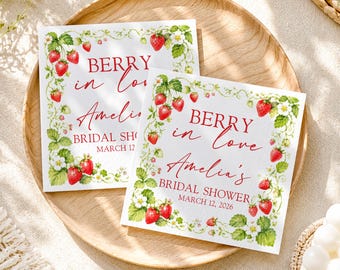 May include: Two square white paper napkins featuring a watercolor strawberry vine border with red berries and green leaves. The text reads BERRY in love Amelia's BRIDAL SHOWER MARCH 12, 2026 in red script and block lettering, centered on the white background.
