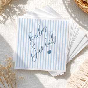 May include: A stack of square white paper napkins featuring a light blue vertical striped pattern. The text Baby Daniel is printed in a dark blue cursive script in the center, accompanied by a small dark blue heart icon below the name.