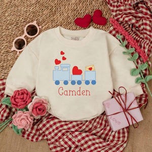 May include: Cream-colored baby bodysuit with a blue train design carrying red hearts and an envelope. The name "Camden" is embroidered in red. The bodysuit is surrounded by Valentine's Day themed decorations.