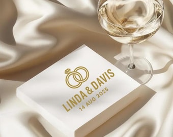 May include: A square white wedding napkin with gold text and a ring graphic. The text reads "LINDA & DAVIS 14 AUG 2025". A glass of champagne sits next to the napkin on a white satin surface.