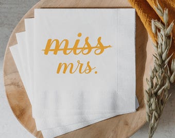 May include: White paper napkins with the words "miss mrs." in a yellow script font. The napkins are stacked on a wooden plate, with a yellow cloth and dried wheat stalks in the background.