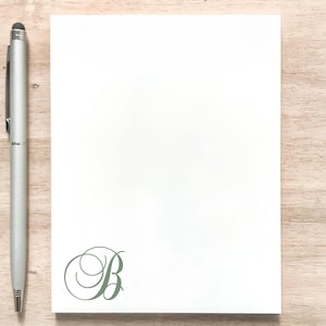 May include: A white notepad with a green, stylized letter "B" in the bottom right corner. A silver pen rests on the left side of the notepad. The notepad and pen are on a light brown wooden surface.