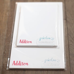 May include: A pack of personalized stationery featuring the name "Addison" in pink script. The stationery is white with a blue and white logo for "Jubilee Design Studio" with the text "Stationery & Invitations" and the website address "www.jubileedesignstudio.com".