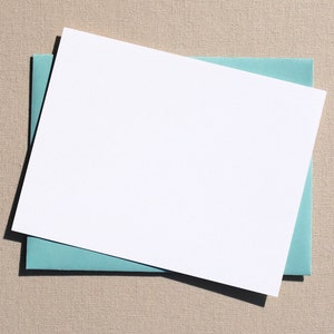 Puede incluir: Una tarjeta blanca con un sobre azul claro debajo. La tarjeta y el sobre est&aacute;n colocados planos sobre una superficie beige texturizada.