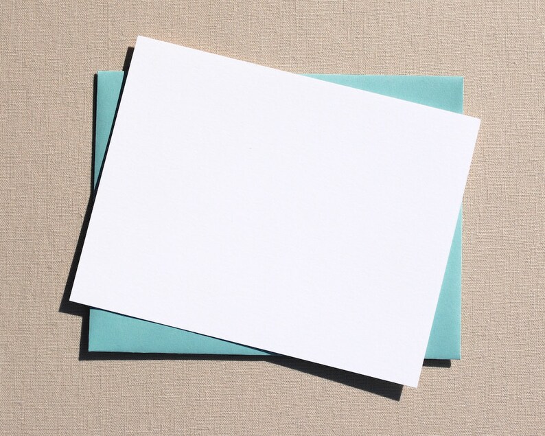 Puede incluir: Una tarjeta blanca con un sobre azul claro debajo. La tarjeta y el sobre est&aacute;n colocados planos sobre una superficie beige texturizada.