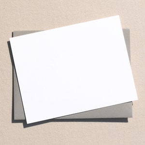 May include: A blank white card with rounded corners is placed on top of a gray envelope. The envelope and card are on a beige textured surface.